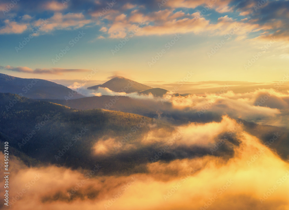 Aerial view of mountains in orange clouds at sunrise in summer. Mountain peak in fog. Beautiful landscape with high rocks, hills, sky. Top view from drone of mountain valley in low clouds. Foggy hills