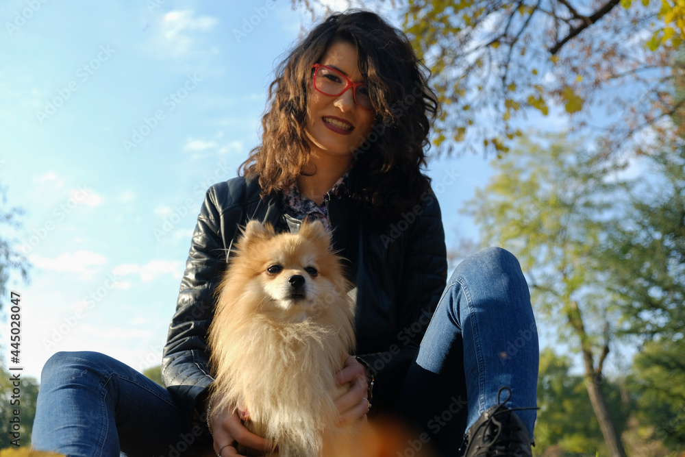 Portrait of a happy woman and her pomeranian pet dog sitting on the ground in a park full of autumn leaves.