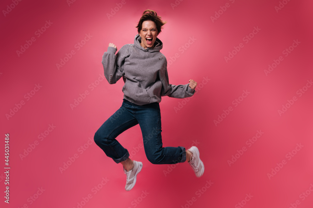 Optimistic woman in jeans and hoodie jumping on pink background. Happy teen girl in denim pants moves on isolated