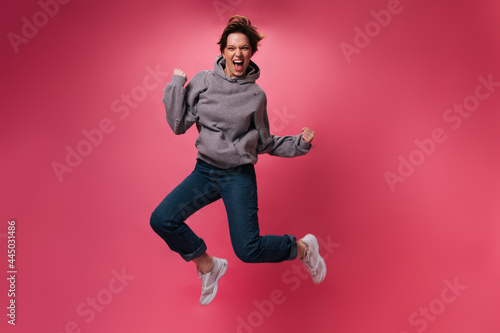 Optimistic woman in jeans and hoodie jumping on pink background. Happy teen girl in denim pants moves on isolated