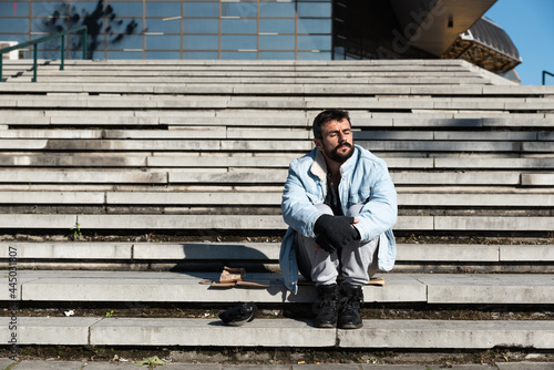 Young sick mentally ill and hungry homeless man sitting on the stairs and sunbathing on the cold sunny day in the urban city street, social documentary concept 