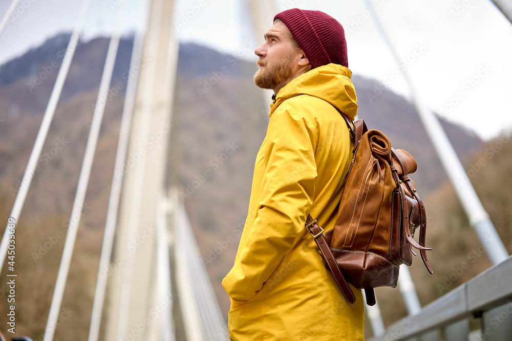 Dreamy caucasian male with beard in casual yellow coat with leather backpack, in hat, looking up, exploring new places, on bridge, copy space. portrait