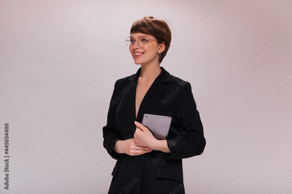 Business woman in black suit holds computer tablet. Short-haired employee in dark jacket smiles widely on isolated backdrop Business woman in black suit holds computer tablet. Short-haired employee in dark jacket smiles widely on isolated backdrop