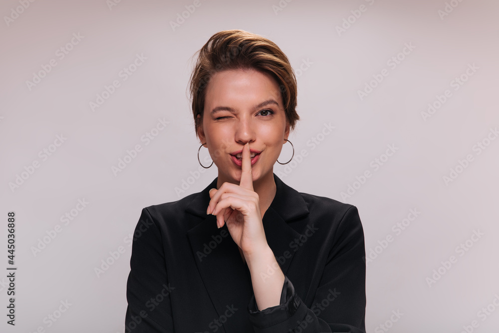 Woman winking and asking for keeping secret. Short-haired lady in stylish jacket posing and looking into camera on isolated Woman winking and asking for keeping secret. Short-haired lady in stylish jacket posing and looking into camera on isolated