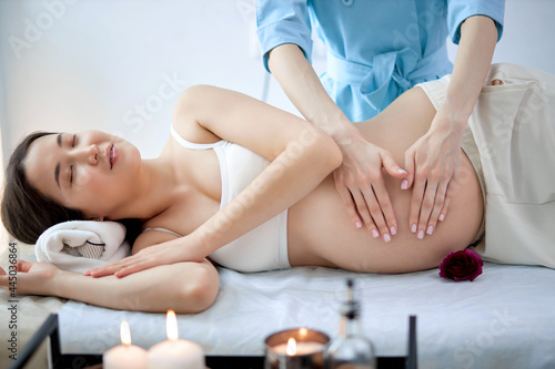 Female therapist hands massaging asian pregnant woman,for treat and relax program,at spa therapy. Side view on masseur in blue uniform massaging gravid female lying on bed in spa cabinet