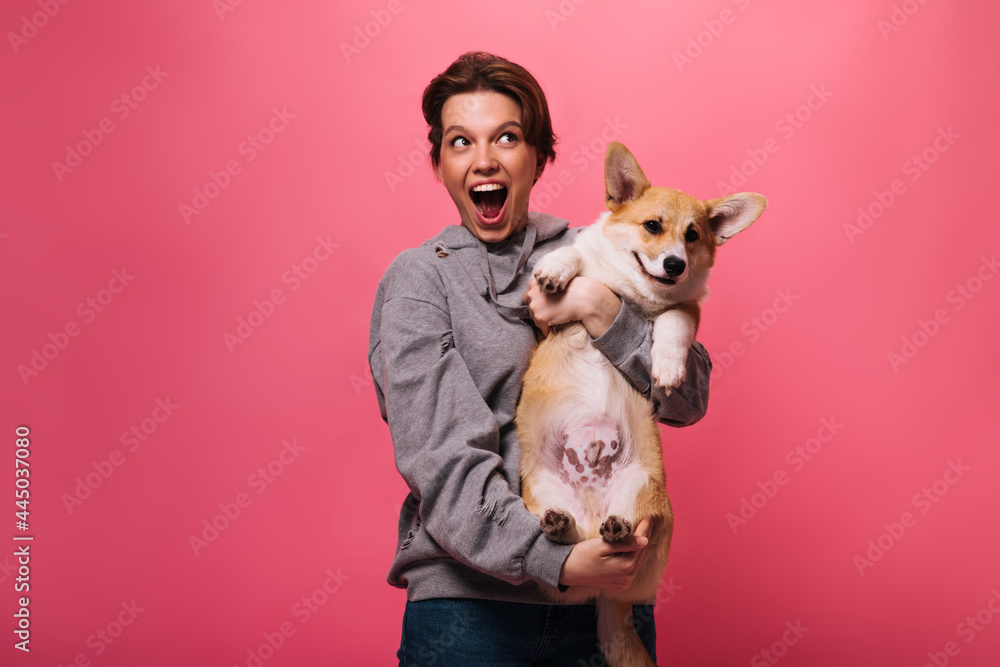 Joyful woman in grey hoodie holding corgi on pink background