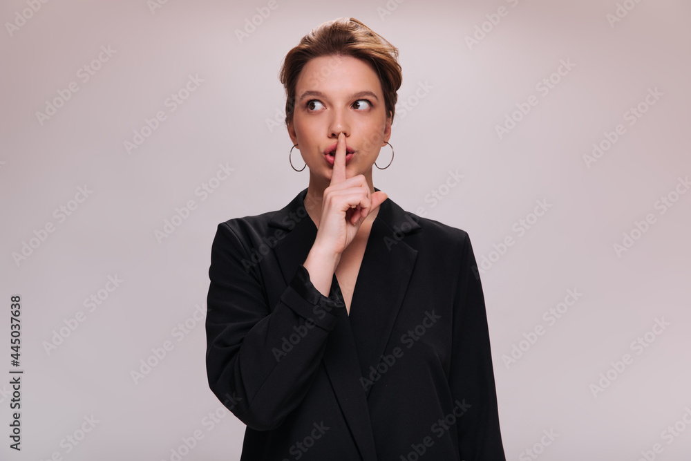 Woman in jacket asks to keep secret. Pretty girl in black suit wants to hear silence. Lady in oversize outfit poses on isolated background Woman in jacket asks to keep secret. Pretty girl in black suit wants to hear silence. Lady in oversize outfit poses on isolated background