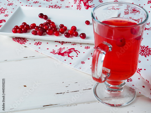 Selective focus on cranberries on a white rectangular ceramic plate. Berry drink in a glass cup. Napkin with Christmas ornament. New Year's decor on a white wooden background. Copy space.