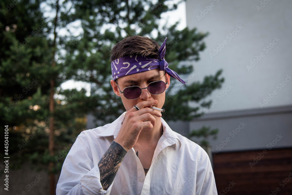 Foto Stock Positive male with bandana dressed in street style clothes with  chains around neck and purple bandana for a walk. Youth and lifestyle  concept. Rap artist new school. Smoking cigarette.
