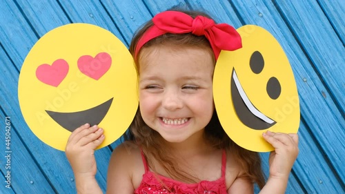 A little girl covers her face with a cardboard smiley face with happy and loving faces . World Emoji Day. An anthropomorphic smiley face.   photo