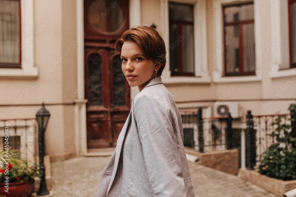 Fototapeta premium Lady in grey jacket looks into camera on background of building. Short-haired woman in oversize suit walks outside