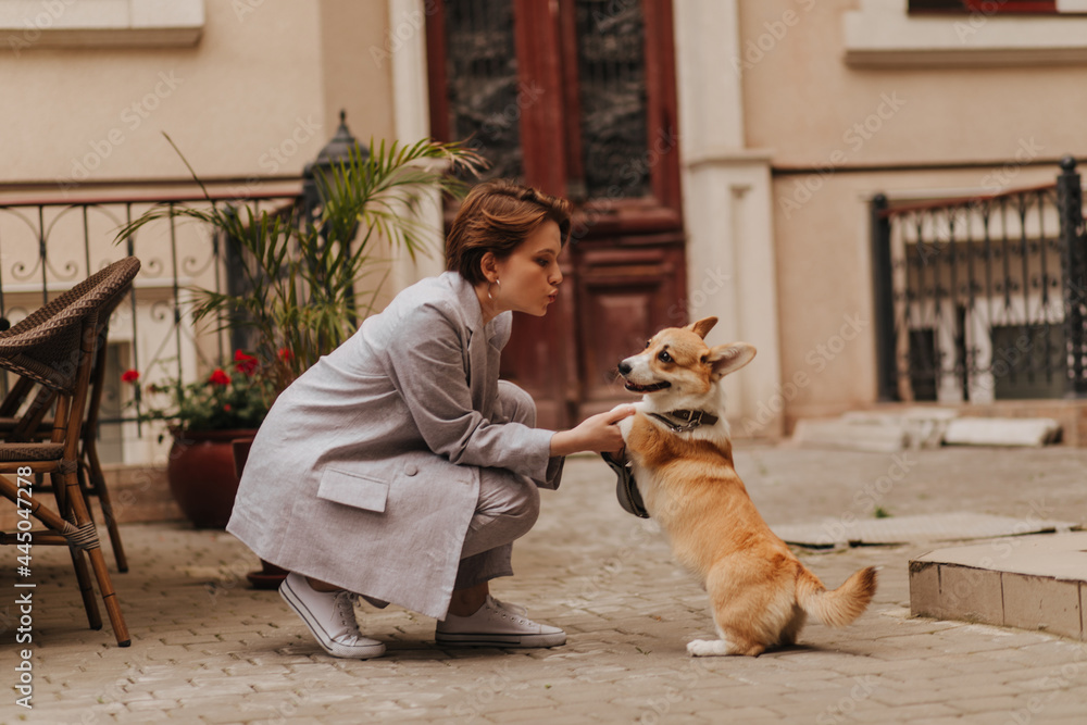 Cool woman in grey outfit plays with corgi outside. Attractive girl in oversize jacket and pants hugging her dog