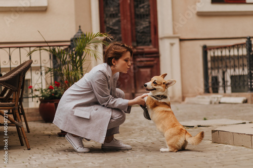 Cool woman in grey outfit plays with corgi outside. Attractive girl in oversize jacket and pants hugging her dog © Look!