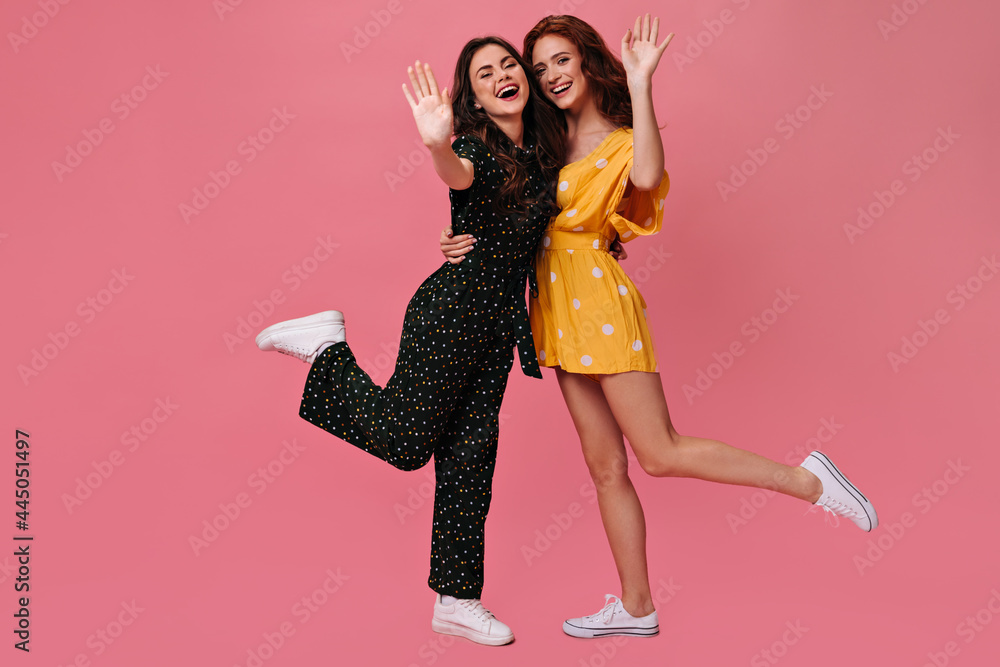 Cheerful women in dresses waving hands in greeting. Two charming girlfriends in beautiful summer clothes rejoice on pink background