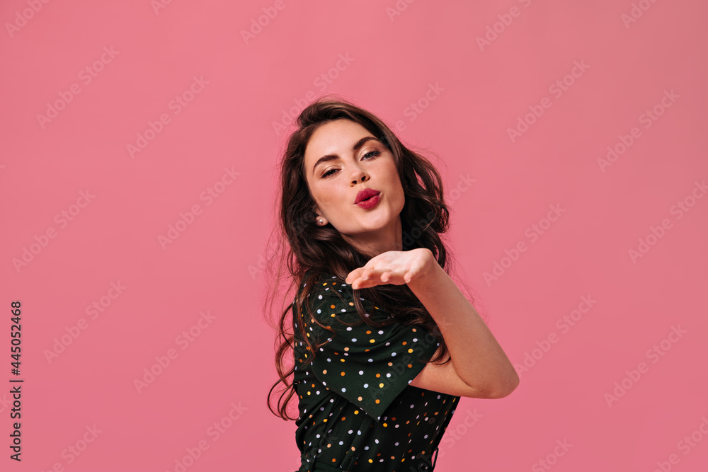 Curly lady with bright lipstick blows kiss on isolated background. Cute girl in dark polka dot dress beautifully and womanly poses in good mood