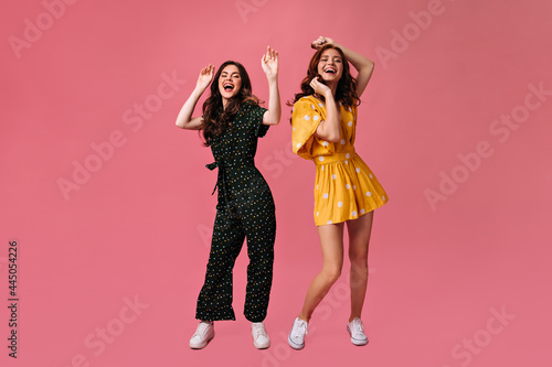 Pretty women in summer stylish outfits dancing on pink background. Beautiful girls in a good mood laugh and move to music