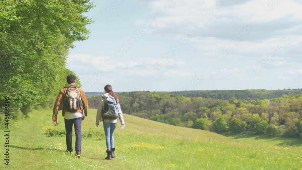 Rear view of couple with backpacks hiking along path across field by forest in countryside - shot in slow motion Rear view of couple with backpacks hiking along path across field by forest in countryside - shot in slow motion