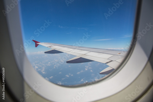 View through a window of an left airplane wing aircraft flying above the clouds with blue sky as travel by airplane concept.