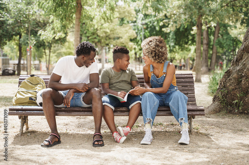 Black family doing school homework sitting on a bench in the park. Little son doing school work with help of his parents