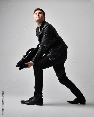 Full length portrait of a  brunette man wearing leather jacket  and holding a science fiction gun.  Standing  action pose isolated  against a grey studio background.