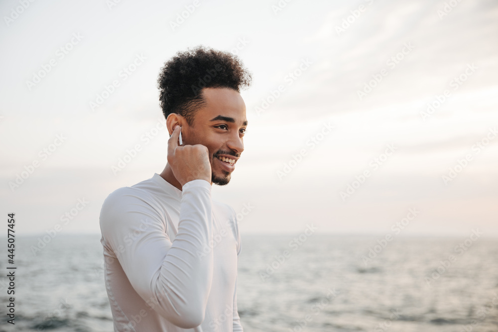 Cheerful young curly brunette dark-skinned man in white t-shirt smiles sincerely and listens to music in wireless headphones near sea.