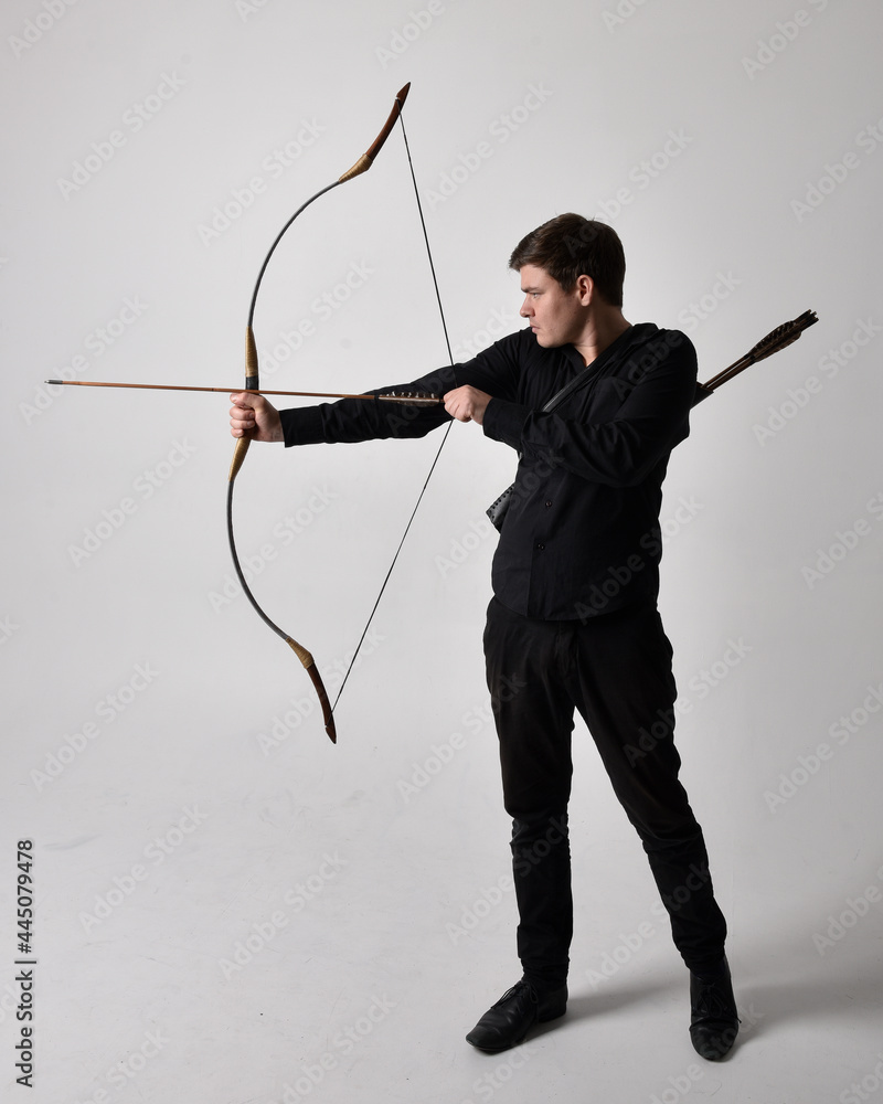 Full length portrait of a  brunette man wearing black shirt and waistcoat holding  a bow and arrow.  Standing  action pose isolated  against a grey studio background.