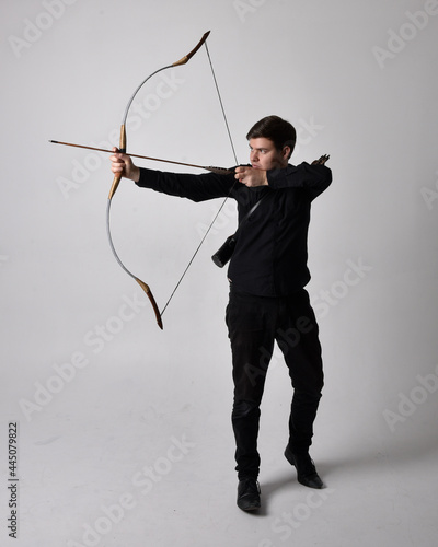 Full length portrait of a brunette man wearing black shirt and waistcoat holding a bow and arrow. Standing action pose isolated against a grey studio background.