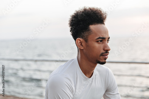 Portrait of curly brunette dark-skinned thoughtful man in white sport long-sleeved t-short looking away near sea.
