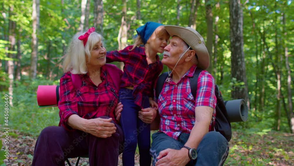 Old senior elderly married family couple resting in forest with granddaughter. Grandmother grandfather and child kid girl tourists sitting in wood, hugging, kissing. Active hiking and tourism outdoors Old senior elderly married family couple resting in forest with granddaughter. Grandmother grandfather and child kid girl tourists sitting in wood, hugging, kissing. Active hiking and tourism outdoors