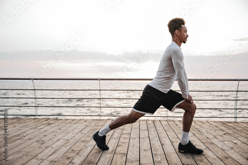 Strong active curly dark-skinned man in black shorts and white long-sleeved t-shirt squats, works out and stretches near sea.