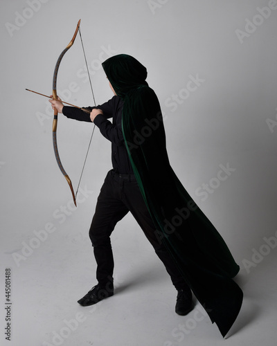 Full length portrait of a  brunette man wearing black shirt, waistcoat and a green velvet cloak holding a bow and arrow.  Standing  action pose isolated  against a grey studio background.