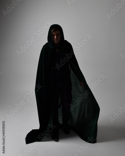Full length portrait of a brunette man wearing black shirt, waistcoat and a green velvet cloak. Standing action pose isolated against a grey studio background.