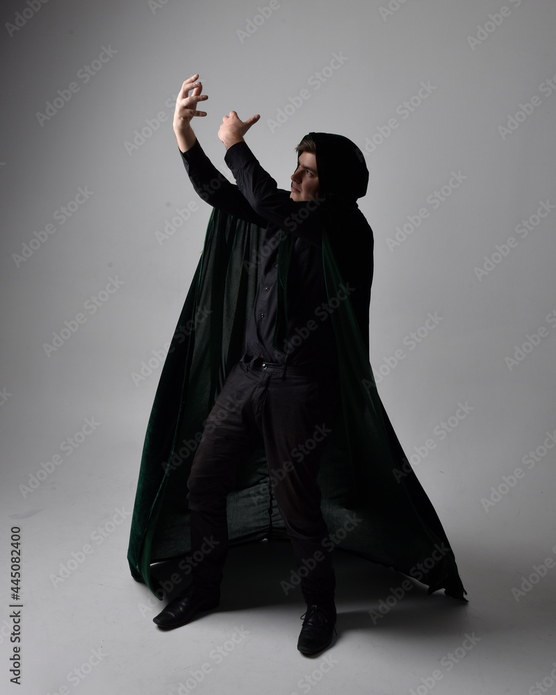 Naklejka premium Full length portrait of a brunette man wearing black shirt, waistcoat and a green velvet cloak. Standing action pose isolated against a grey studio background.