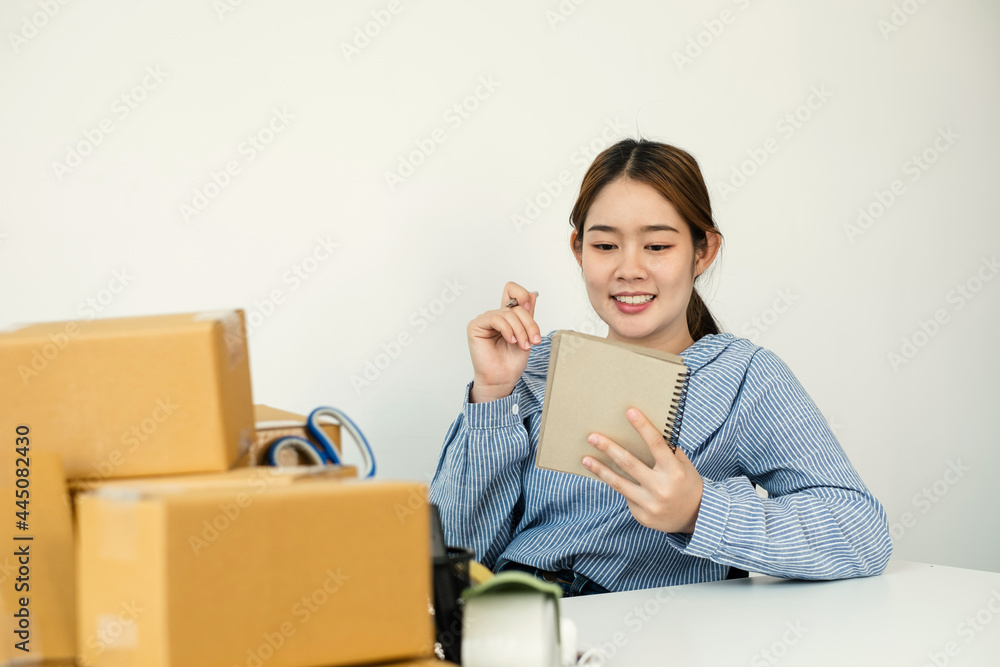 Young Asian woman business owner with many parcel boxes on the table happy online sales job, use the laptop, get an order from customers, take notes, and make arrangements for delivery by post Young Asian woman business owner with many parcel boxes on the table happy online sales job, use the laptop, get an order from customers, take notes, and make arrangements for delivery by post