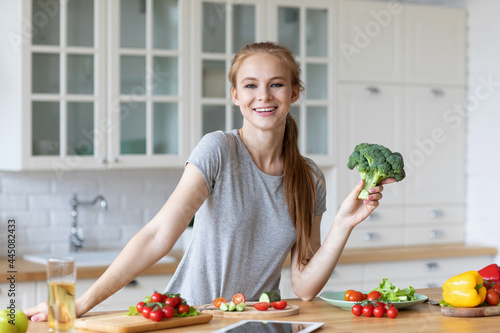 Young happy woman holding broccoli in kitchen with green fresh ingredients indoors. Healthy food