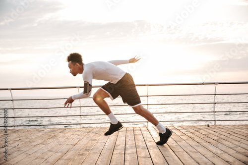 Young dark-skinned man in white long-sleeved t-shirt and black shorts running in terrace near sea. Sportsman working out outdoors.