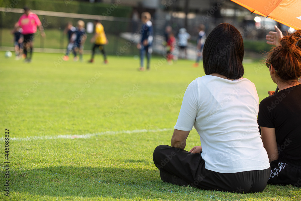 Moms sitting and watching their sons playing football in a school tournament on a sideline with a sunny day. Sport, outdoor active, lifestyle, happy family and soccer mom and soccer dad concepts. Moms sitting and watching their sons playing football in a school tournament on a sideline with a sunny day. Sport, outdoor active, lifestyle, happy family and soccer mom and soccer dad concepts.