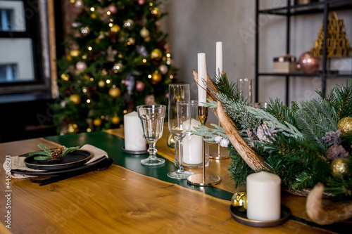 Christmas table setting in rustic style with candles, plates, glasses and eco decors on wooden festive table.New year decorations with fir branches, golden balls, pine cones. Cozy winter holidays.