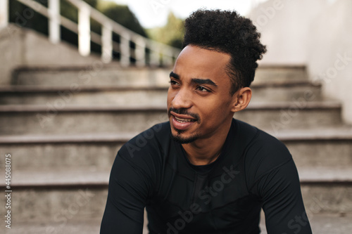 Charming brunet curly dark-skinned man in sport long-sleeved black t-shirt looks away  smiles gently and poses near stairs.