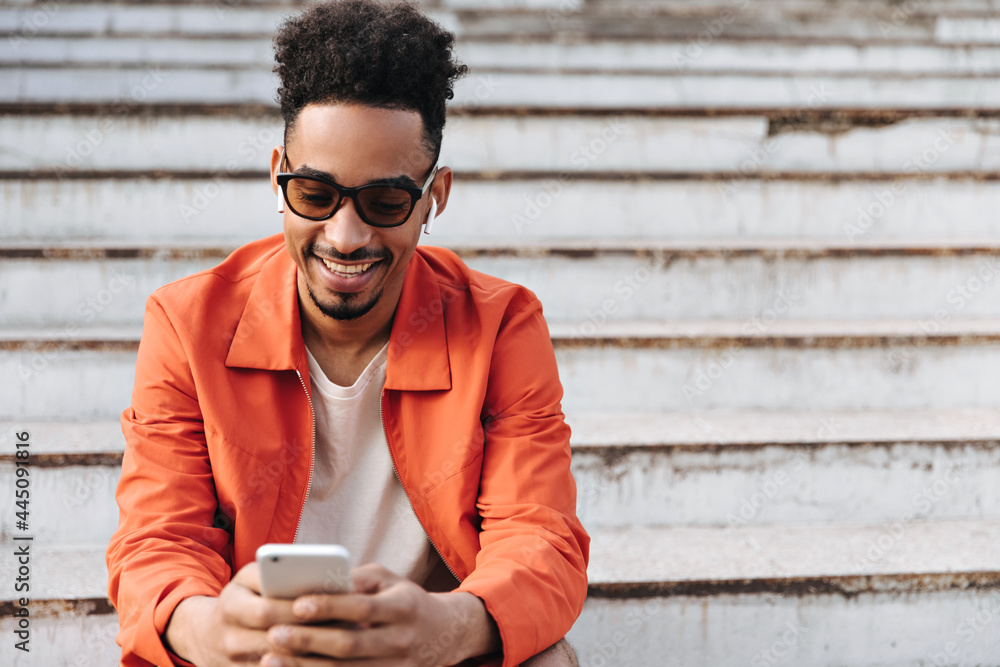Fototapeta premium Excited young charming bearded man in sunglasses and orange jacket smiles sincerely, sits on stairs and holds phone outside.