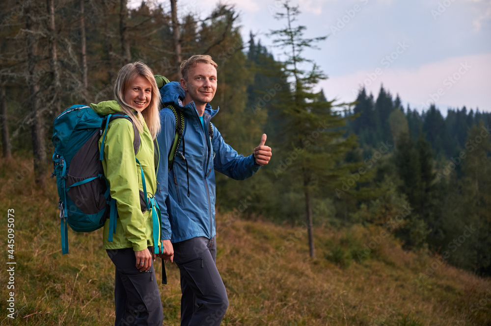 Two young tourists with joyful facial expressions holding hands while walking outdoors. Man pleased with hike at mountain trails and lawns showing thumb up. Concept of travelling and hiking.