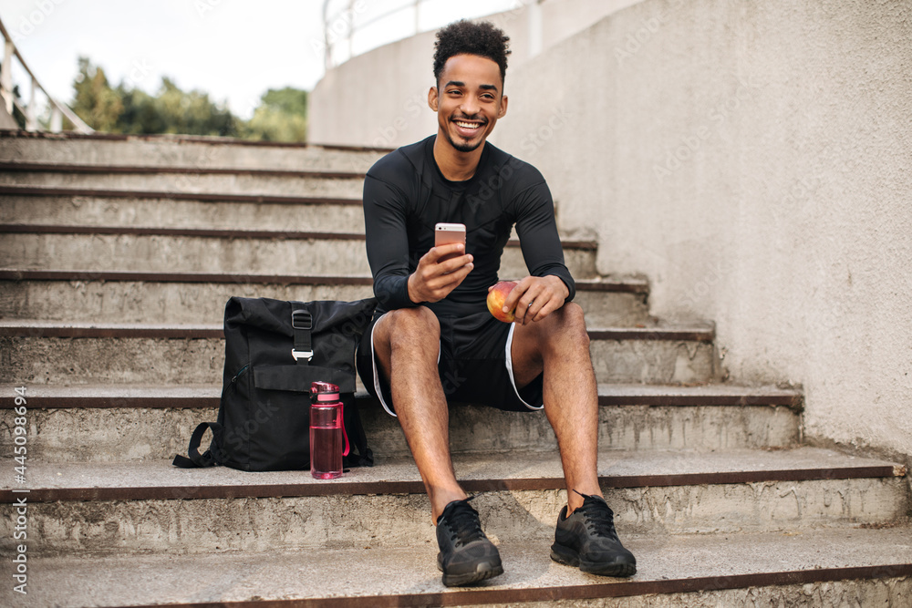 Cool young curly bearded dark-skinned man in black shorts and long-sleeved t-shirt smiles, sits on stairs outside and holds phone. Cool young curly bearded dark-skinned man in black shorts and long-sleeved t-shirt smiles, sits on stairs outside and holds phone.
