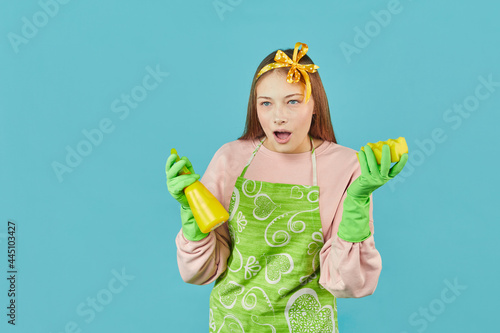 Ready For Spring-Cleaning. Portrait of joyful girl with household supplies in hands over blue background. Young woman hold household cleaning product and spray bottles.