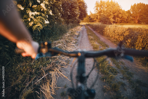 first-person view of riding a bike in a rural area. Cycling through the countryside road on the warm summer evening. Blurred bicycle handlebar with green grass and bushes in the background.