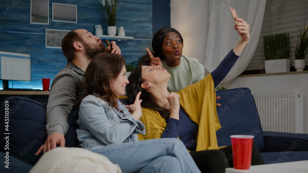 Fototapeta premium Excited group of multiracial friends sitting on sofa during joyful party taking selfie posting on social media. Multi-ethnic friends enjoying time together sharing photos late at night