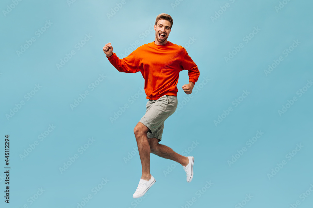 Full length photo of emotional young guy in orange sweatshirt, beige shorts and white sneakers jumping and smiling on isolated backdrop..
