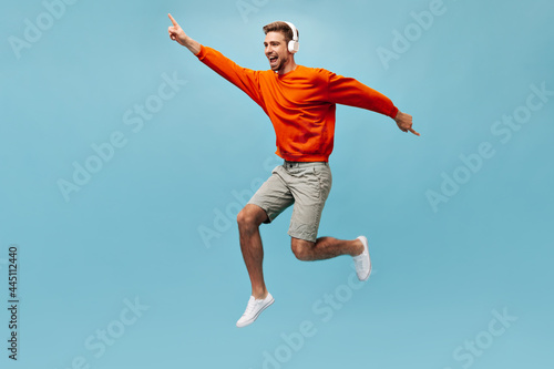 Cheerful modern man with ginger beard and white headphones in stylish sweatshirt and light sneakers jumping and smiling..