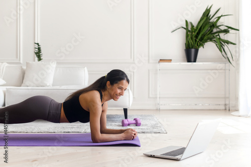 Attractive indian young woman using a laptop for watching sports video tutorial, doing plank exercises in light modern living room, serene athletic mixed-race lady doing workout at home