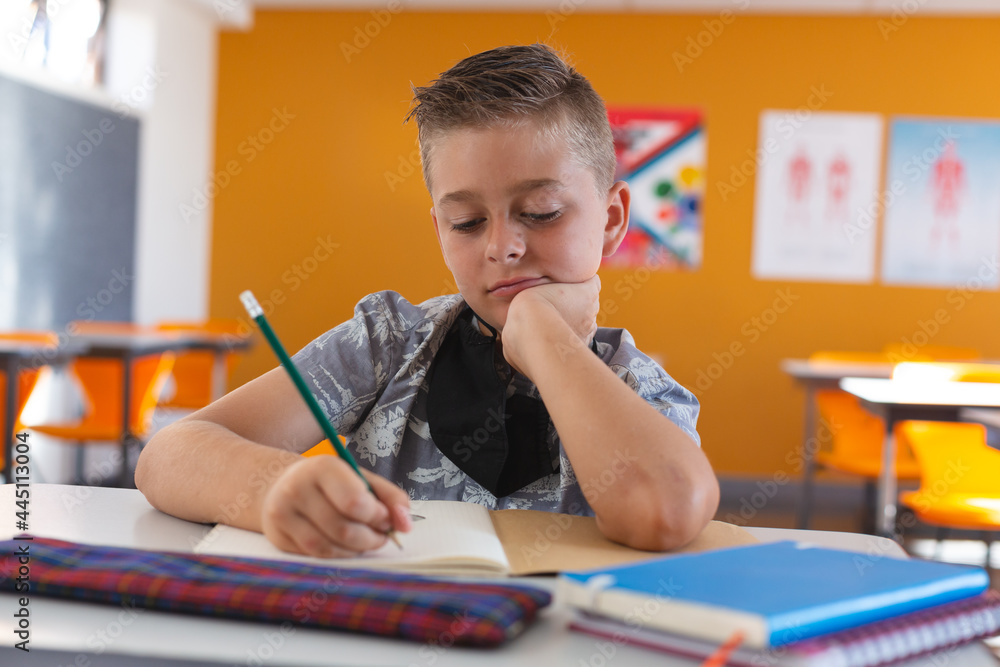 Fototapeta premium Caucasian schoolboy with face mask sitting in classroom concentrating and writing in schoolbook