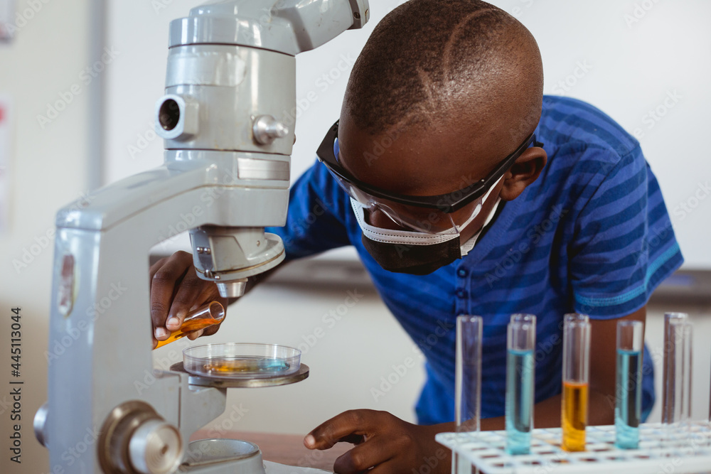 Fototapeta premium African american schoolboy in safety glasses and face mask using microscope in science class